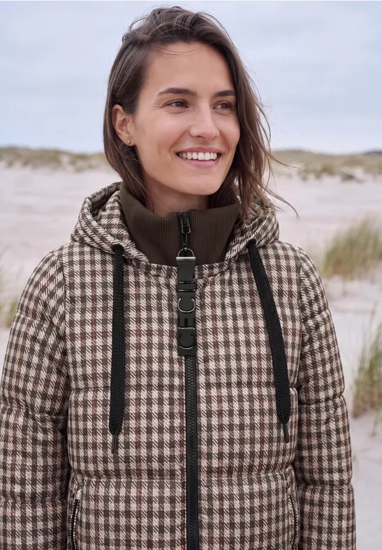 Een glimlachende vrouw in een pied-de-poule jas op het strand voor duinen.