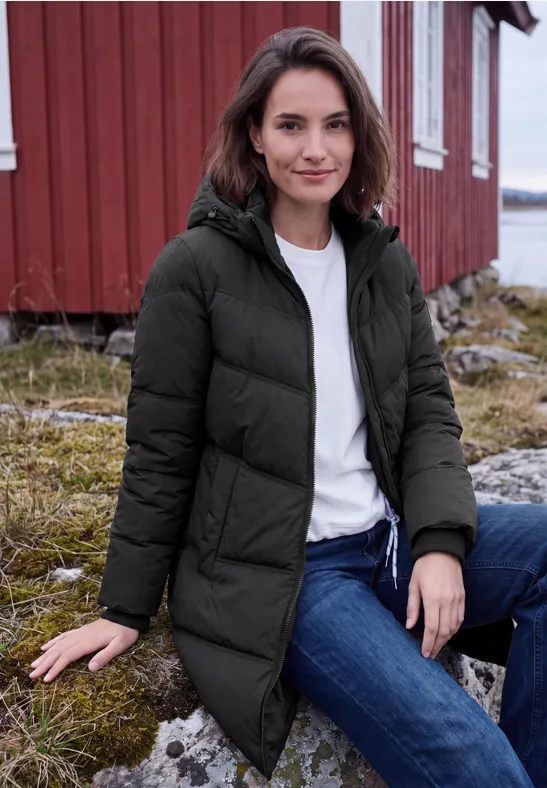 Frau in schwarzer Winterjacke sitzt vor rotem Holzhaus an Küste, trägt weiße Bluse und Jeans.