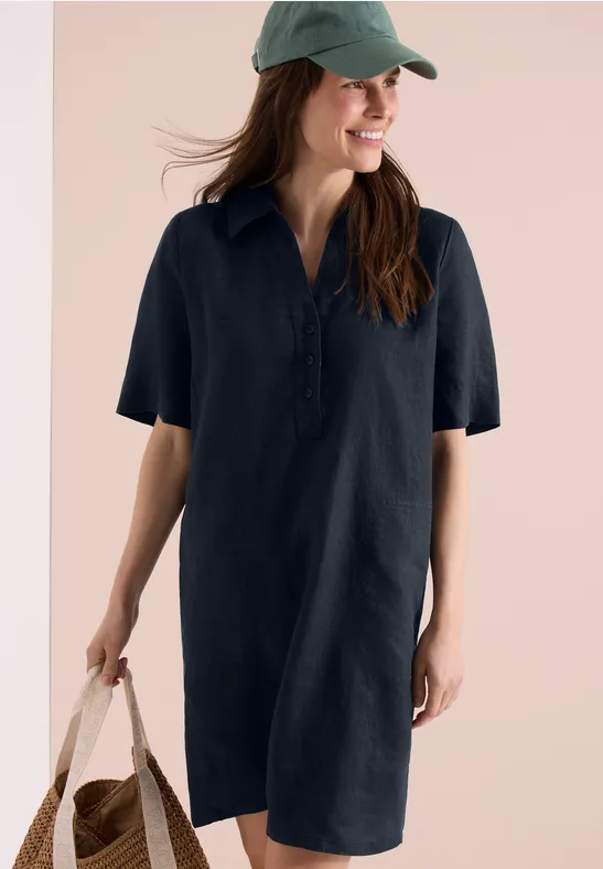 Frau trägt ein locker sitzendes, dunkelblaues Hemdkleid, grüne Cap und hält eine Strohtasche.