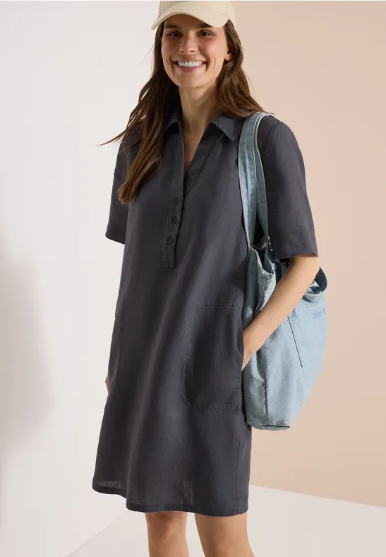 Frau in dunkelgrauem Hemdkleid mit halblangen Ärmeln und hellblauer Schultertasche, Hände in den Taschen.