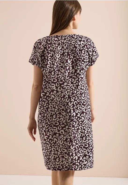 Frau von hinten in knieumspielendem, kurzärmeligem Kleid mit abstraktem Tiermuster vor neutralem Hintergrund.