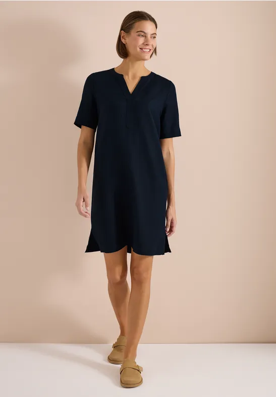 Frau in schlichtem dunkelblauem Etuikleid und beigen Slippers vor neutralem Hintergrund.