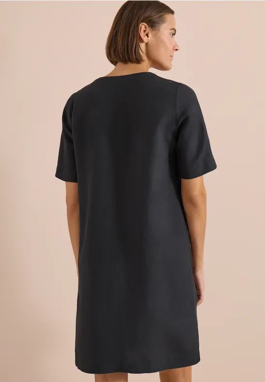 Frau von hinten in einfach geschnittenem, knielangem schwarzen Leinenkleid vor neutralem Hintergrund.