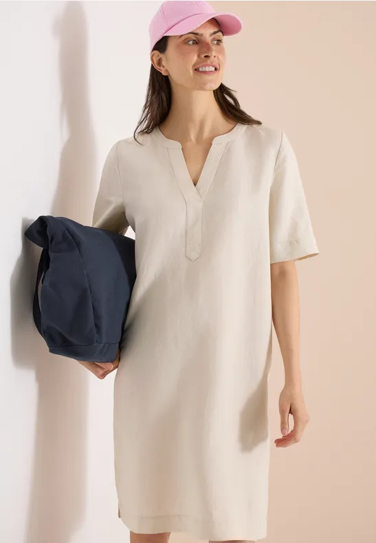 Frau im cremefarbenen Kurzarmkleid mit V-Ausschnitt hält dunkelblaue Tasche und trägt rosa Cap.