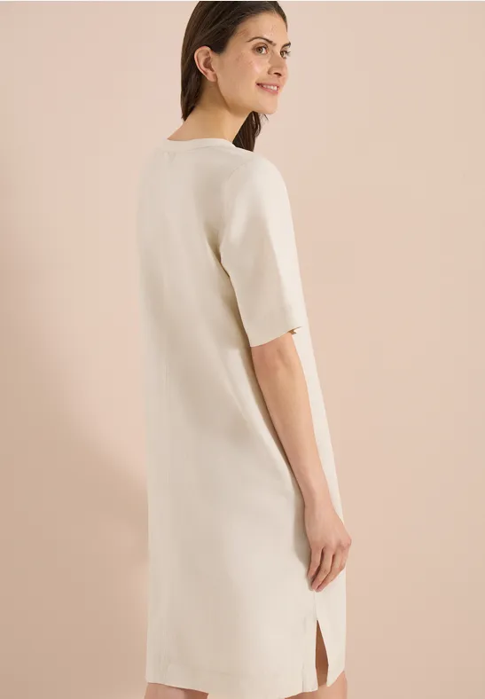 Rückenansicht einer Frau in einem schlichten, cremefarbenen Midi-Kleid mit kurzen Ärmeln und seitlichem Schlitz.