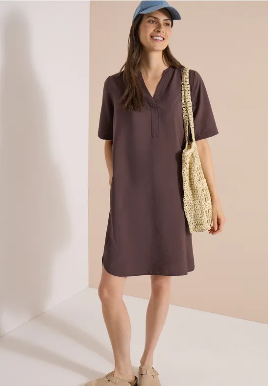 Vrouw in chocoladebruin polojurkje met strooien shopper, staand voor een beige achtergrond.