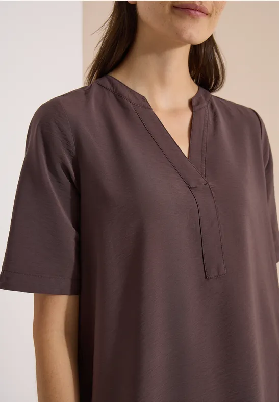 Vrouw draagt een simpele, korte mouw, bruine blouse met V-hals en rechte pasvorm tegen een lichte achtergrond.