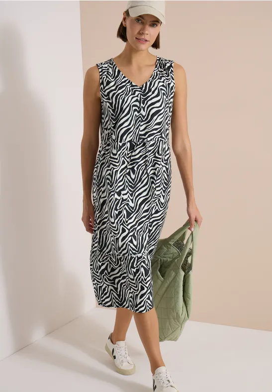 Femme en robe midi zèbrée, baskets blanches et sac vert, look décontracté et moderne.