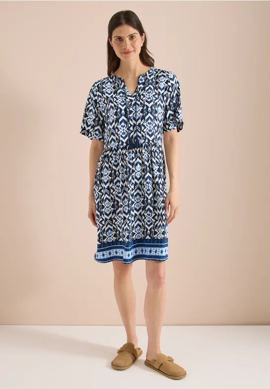 Frau trägt blau-weiß gemustertes knielanges Kleid mit kurzen Ärmeln und Taillengürtel, dazu beigefarbene Slipper.