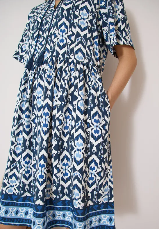 Frau trägt ein langes blau-weiß gemustertes Kleid mit kurzen Ärmeln und Seitentaschen vor hellem Hintergrund.