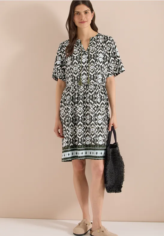 Frau in knielangem schwarz-weiß gemustertem Kleid mit grünem Saum, V-Ausschnitt und kurzen Ärmeln, trägt schwarze Tasche.