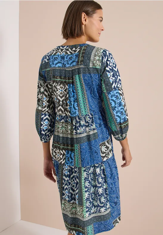Frau in blau-grüner Patchwork-Kleid, 3/4-Ärmel, Rückenansicht vor heller Wand.