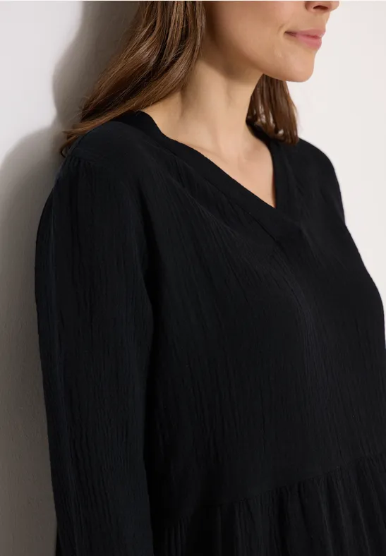 Eine Frau in Nahaufnahme trägt eine schwarze, leicht strukturierte Bluse mit V-Ausschnitt vor heller Wand.