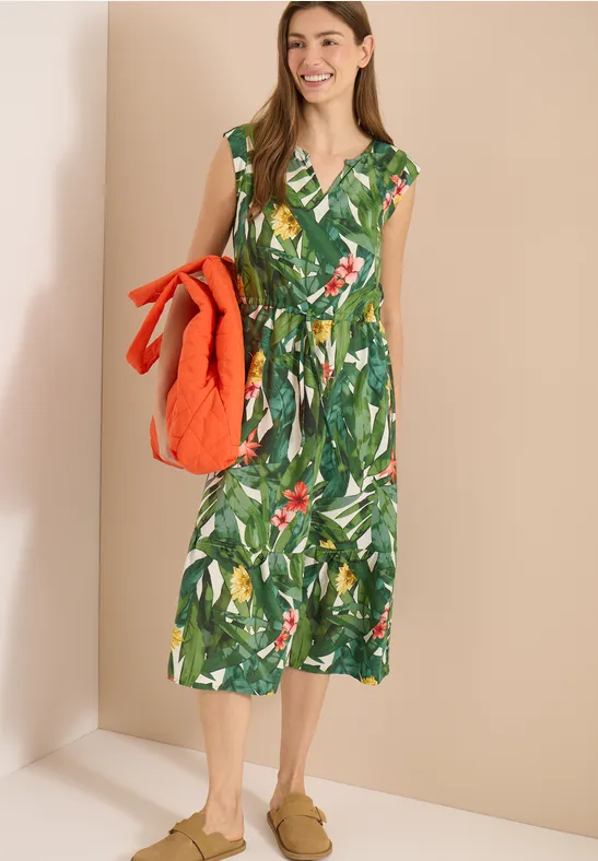Frau in knielangem grünen Kleid mit tropischem Blattprint und V-Ausschnitt, hält eine orangefarbene Steppjacke.
