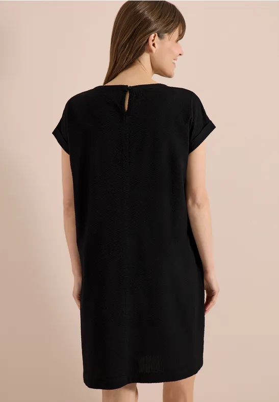 Vrouw van achteren in een zwarte, losvallende jurk met korte mouwen tegen een neutrale achtergrond.