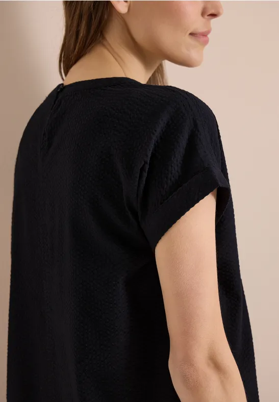 Vrouw van opzij/achterkant in eenvoudige zwarte textuur top met korte mouwen, voor een neutrale achtergrond.