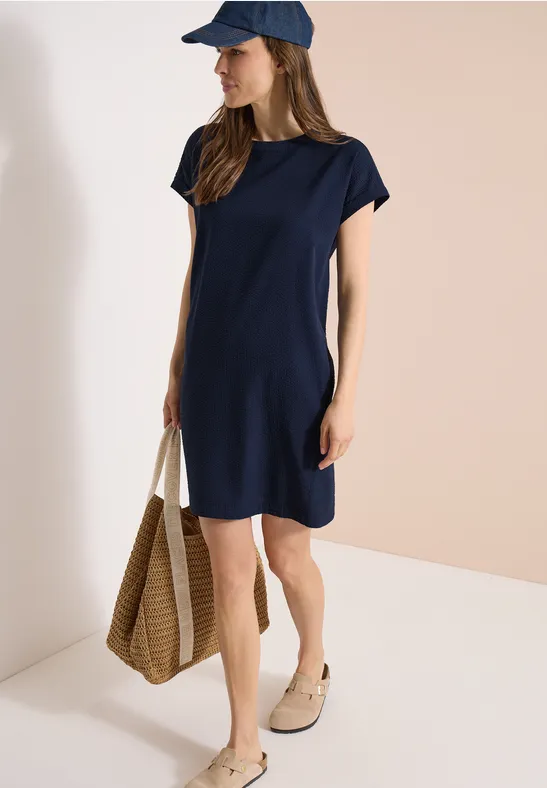 Frau trägt ein schlichtes, navyblaues T-Shirt-Kleid, Kappenärmel, kombiniert mit Cap und Strohtasche.