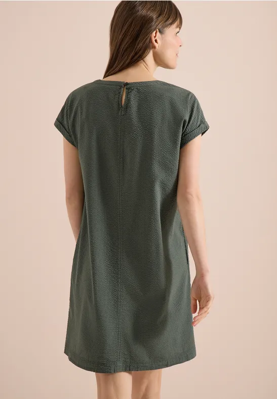 Frau mit Rücken zur Kamera in schlichtem, locker sitzendem, kurzärmligem, dunkelgrünem Kleid.