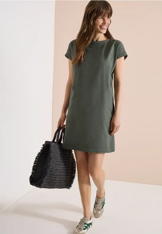 Frau in schlichtem, knielangem olivgrünen Kleid, kurzen Ärmeln, grünen Loafers und großer schwarzer Korbtasche.