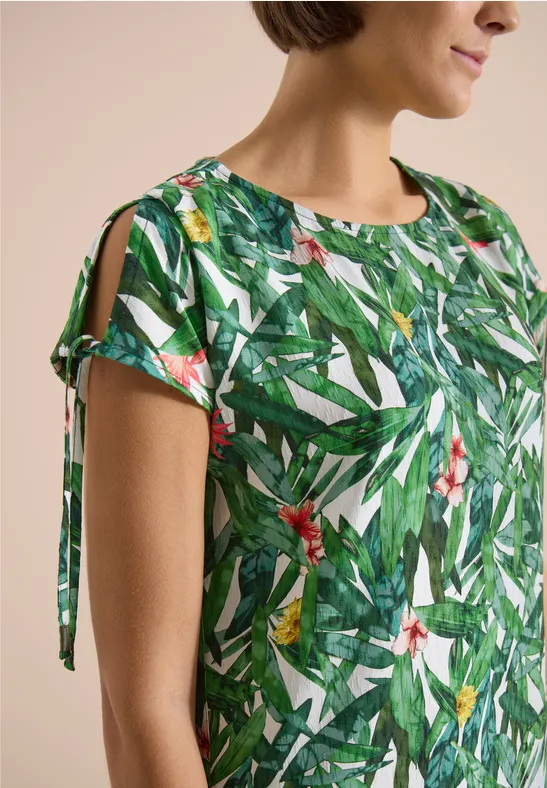 Naheaufnahme einer Frau in grüner Bluse mit tropischem Blatt- und Blumenprint und Ärmeln mit Bändchen.
