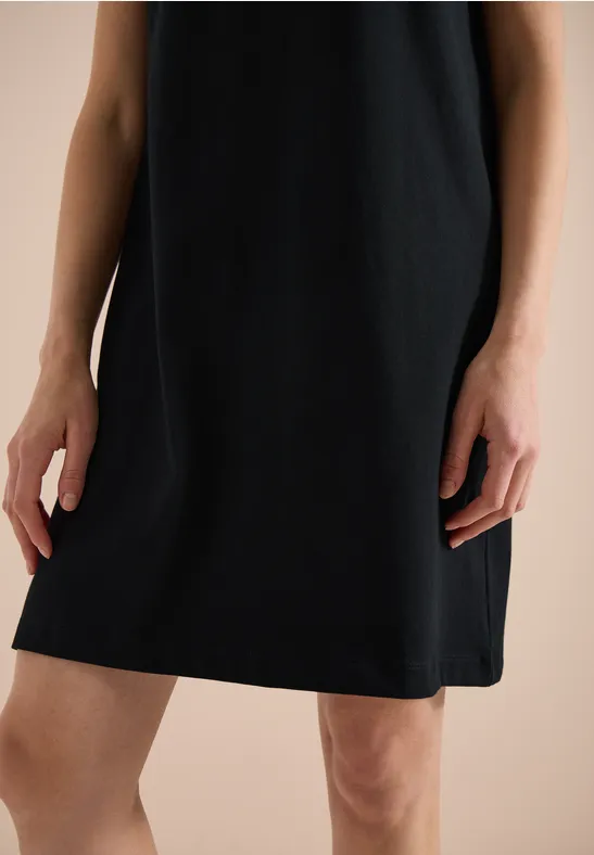 Eine Person trägt ein schlichtes schwarzes Kleid, nur Unterkörper und Hände sind sichtbar.