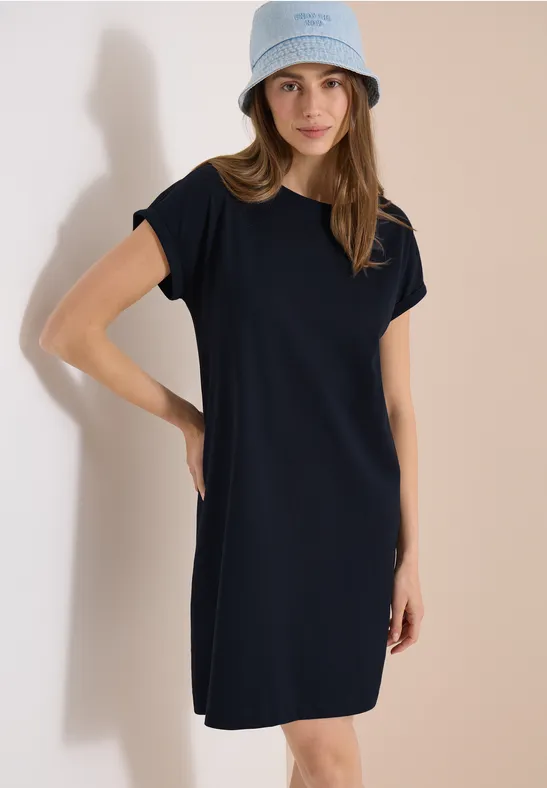 Een vrouw draagt een marineblauwe T-shirtjurk en lichte bucket hat voor neutrale achtergrond.