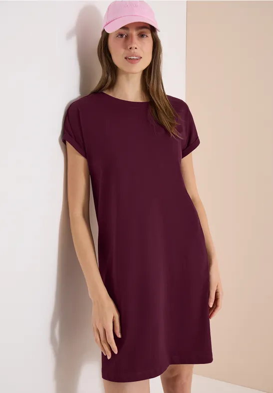 Frau lehnt an einer hellen Wand und trägt ein lässiges, kurzärmeliges, weinrotes T-Shirt-Kleid.