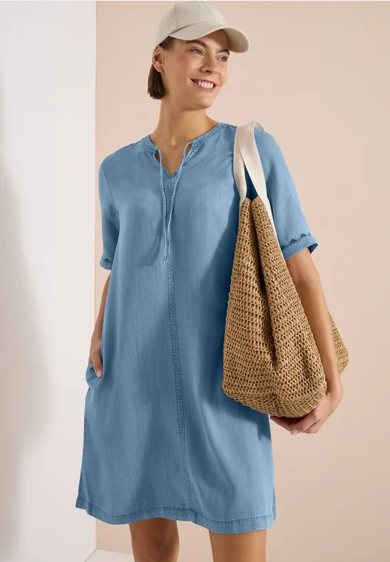 Vrouw in lichtblauw losvallend overhemdjurk met opgerolde mouwen, draagt rieten tas en pet