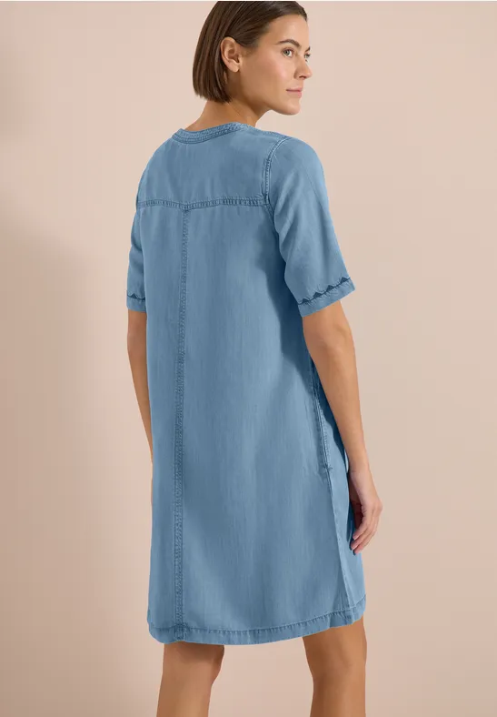 Achteraanzicht van een vrouw in een loszittende blauwe spijkerjurk met halflange mouwen voor een neutrale achtergrond.