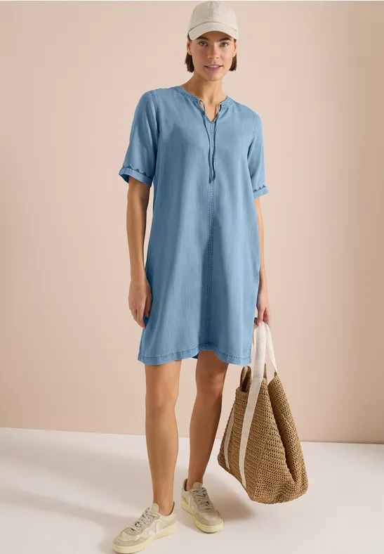 Vrouw in luchtig lichtblauw jurkje met witte sneakers en rieten tas, staand voor een neutrale achtergrond.