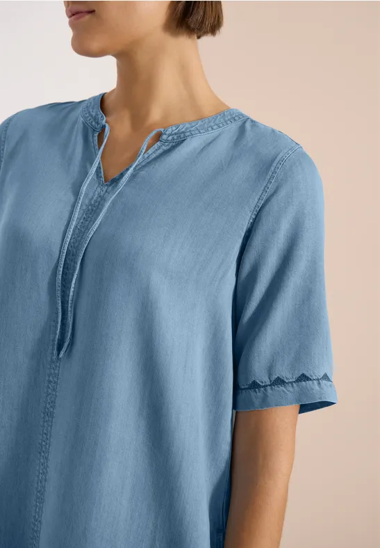 Close-up van een lichtblauwe damesblouse met V-hals en sierstiksel aan de mouwen tegen een beige achtergrond.