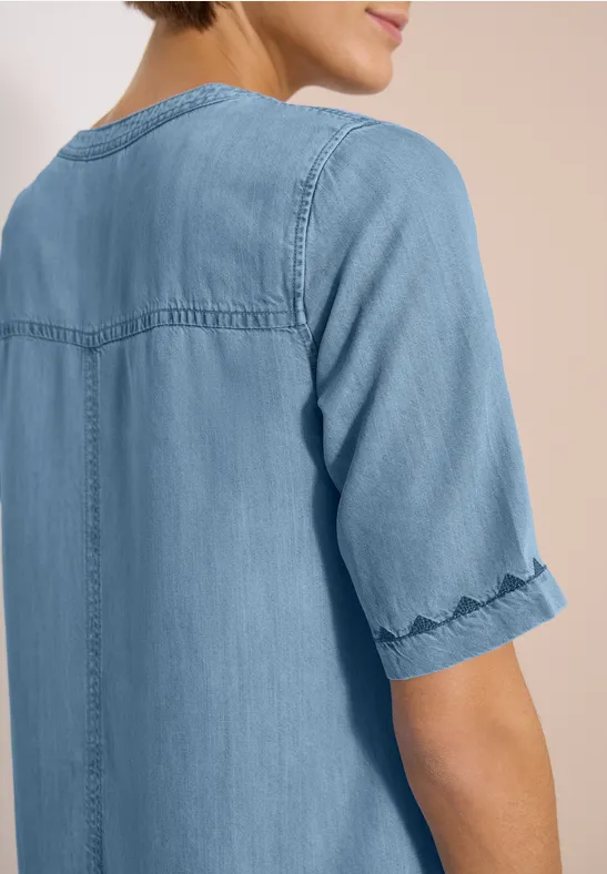 Rugaanzicht van een persoon in een lichtblauw denim top met halflange mouwen en siernaden.