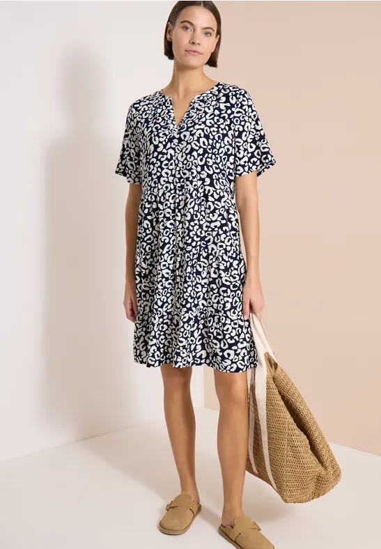 Frau trägt kurzärmeliges, blau-weiß gemustertes Sommerkleid, kombiniert mit Basttasche und beigen Slippern.