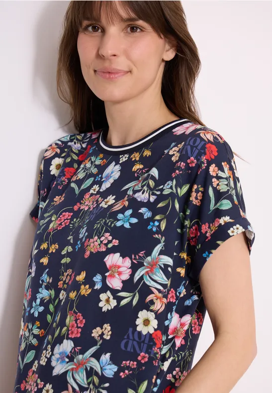 Frau trägt dunkelblaues T-Shirt mit buntem Blumenmuster und kurzen Ärmeln, lächelnd.