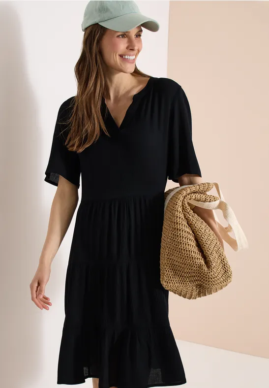 Frau in schwarzem Stufenkleid mit V-Ausschnitt, trägt eine Kappe und hält eine geflochtene Strohtasche.