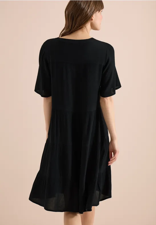 Dame in locker sitzendem, knielangem, schwarzem Kleid mit kurzen Ärmeln, fließendem Stoff, Rückenansicht