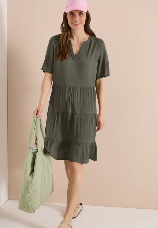 Frau in lockerem, olivgrünem Stufenkleid hält eine gesteppte, hellgrüne Tasche.