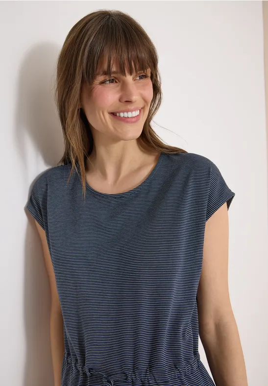 Eine schlanke Frau steht vor weißer Wand und trägt ein dunkelblaues T-Shirt.