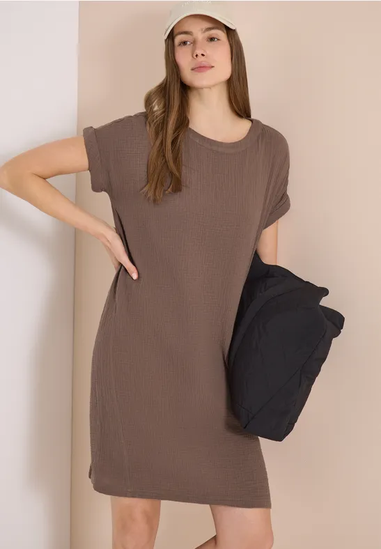 Frau in einem locker sitzenden, dunkelbraunen T-Shirt-Kleid hält lässig eine schwarze Jacke seitlich.