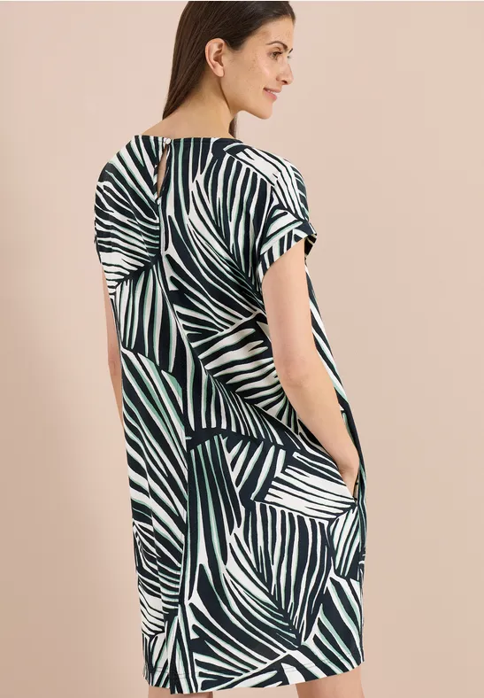 Frau vor hellem Hintergrund in locker fallendem Kurzarmkleid mit grün-weißem Blattmuster und seitlichen Taschen.