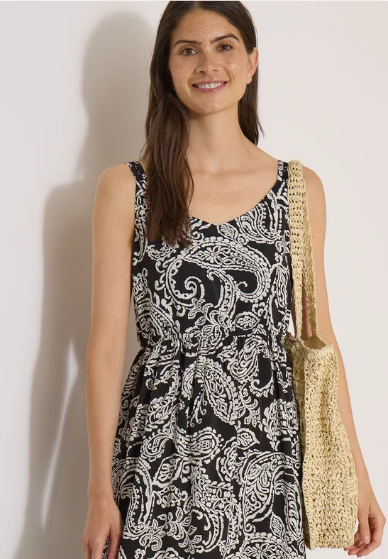 Femme porte une robe d'été sans manches noire à motif paisley blanc et un sac en paille doré.