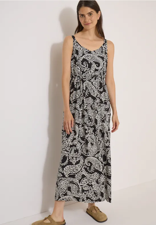 Une femme porte une robe longue sans manches noir et blanc à motif cachemire, avec des mules vert olive.