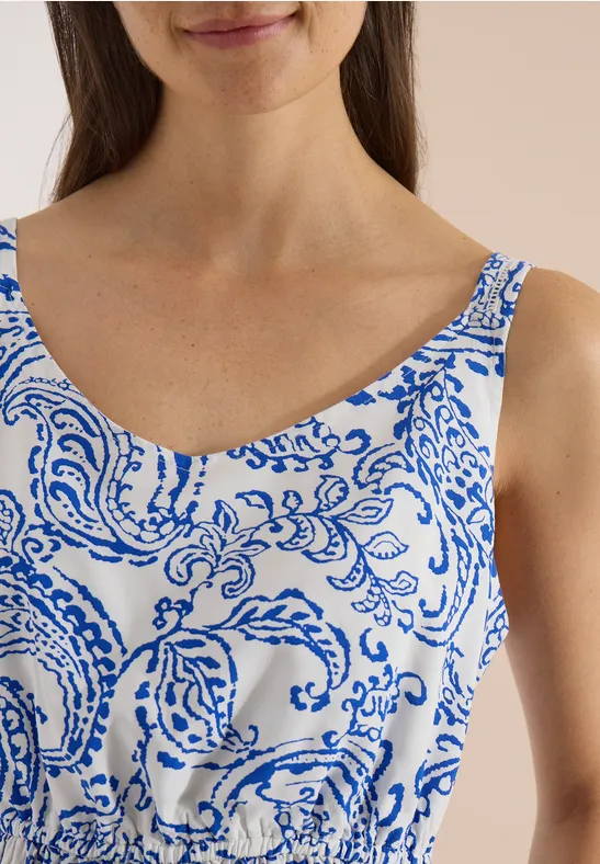 Une femme porte un haut sans manches blanc à motifs bleus ornementaux et à encolure arrondie.