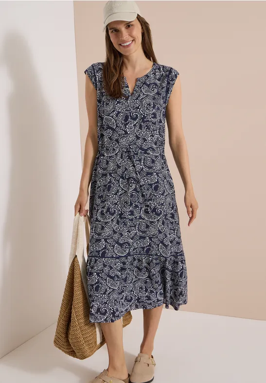 Model trägt ärmelloses Midikleid mit blau-weißem Paisleymuster, V-Ausschnitt, Korbtasche und Sandalen.