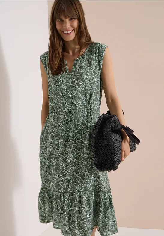 Frau trägt ärmelloses, grünes Maxikleid mit Blumenmuster und Taillengürtel, hält eine runde Strohtasche.