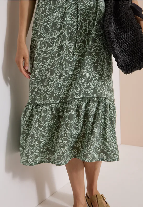 Grünes Midikleid mit Paisley-Muster, Rüschensaum und geflochtenem Handtasche, dazu Sandalen.