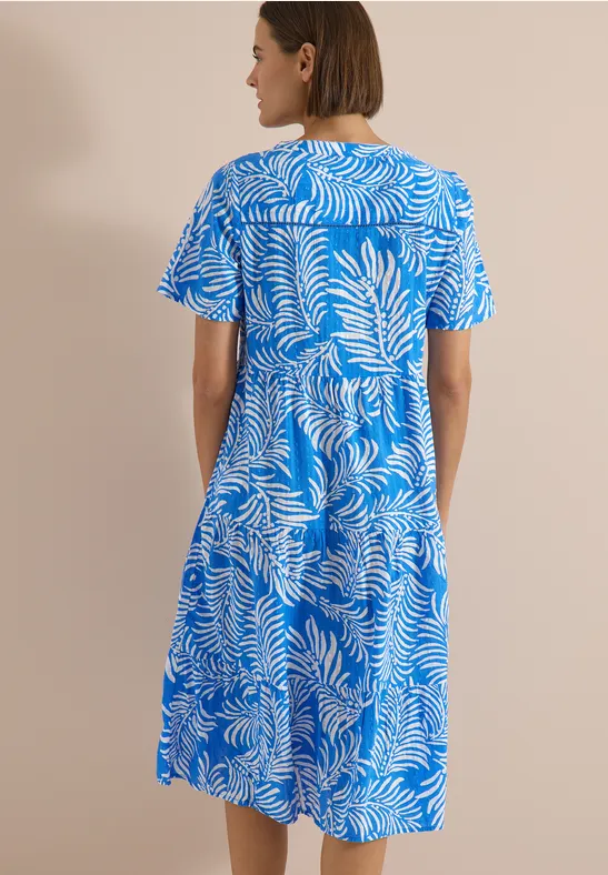 Eine Frau steht mit dem Rücken zur Kamera und trägt ein blau-weißes, kurzärmeliges, knielanges Kleid mit Blattmuster.