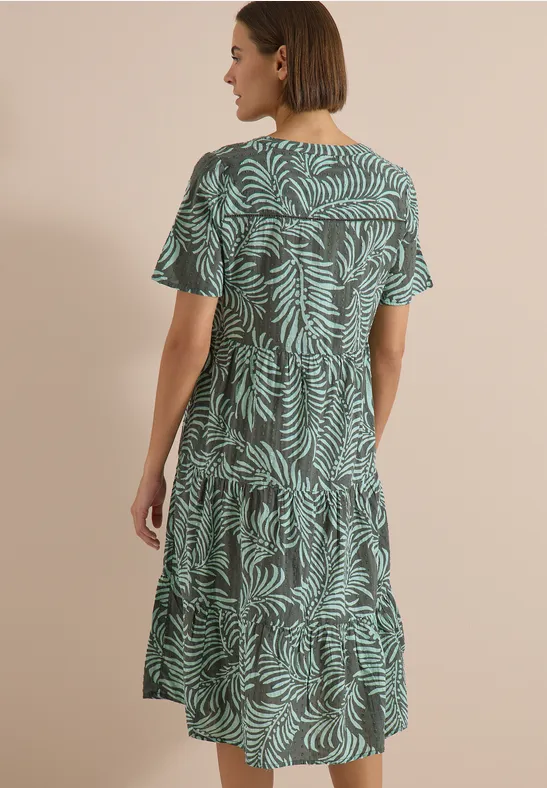 Eine Frau steht mit dem Rücken zur Kamera, trägt ein knielanges, locker sitzendes Kleid mit grünem Blattmuster.