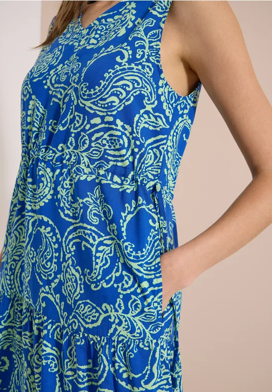Vrouw in zijaanzicht met een mouwloze, blauwe jurk met paisleyprint, hand in de zak.