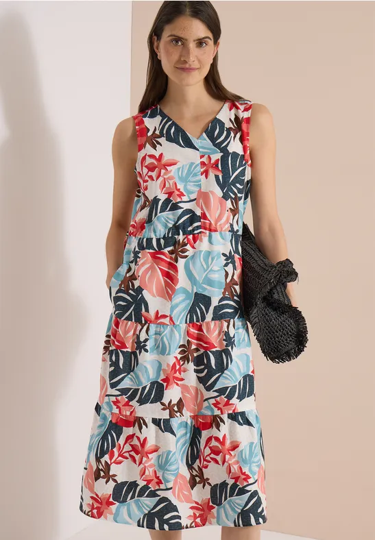 Vrouw in mouwloze zomerse jurk met lagen en kleurrijk bloemen- en bladmotief, met een zwarte gehaakte tas.